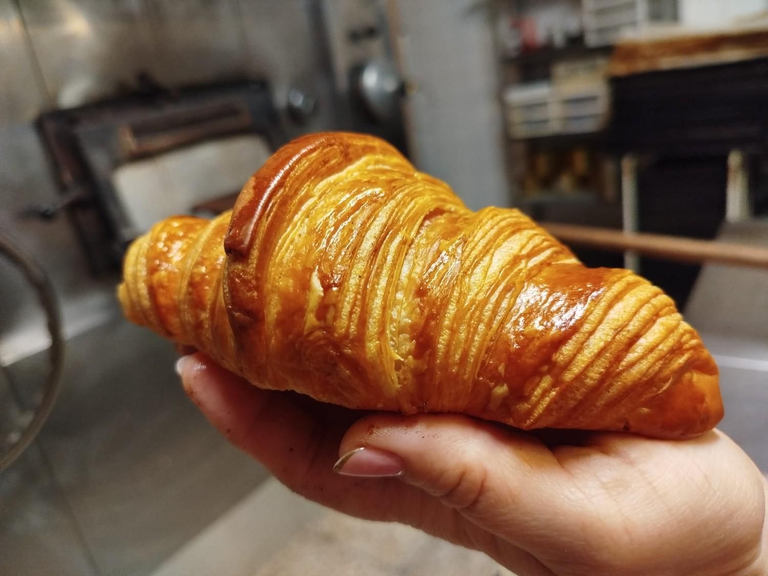 Primer plano de la mano de una persona sosteniendo un cruasán hojaldrado de color marrón dorado en una panadería.
