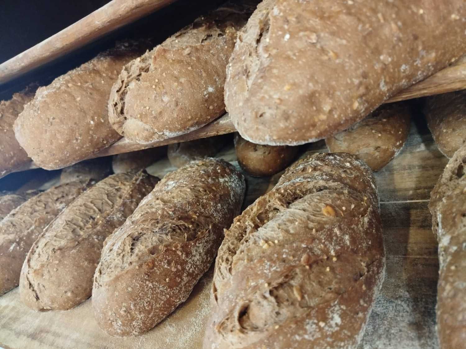 Varias hogazas de pan oscuro, crujiente y con semillas reposan sobre estantes de madera de la panadería.