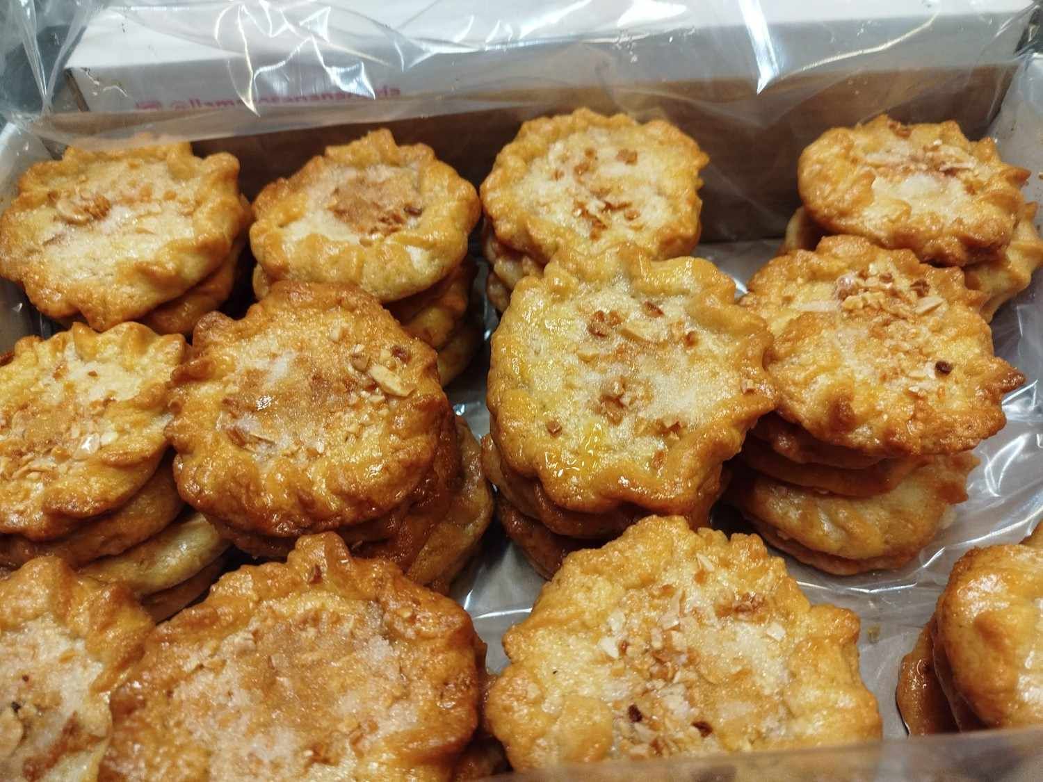 Una caja llena de galletas doradas con bordes festoneados, cubiertas con pequeños trozos de nueces esparcidos.