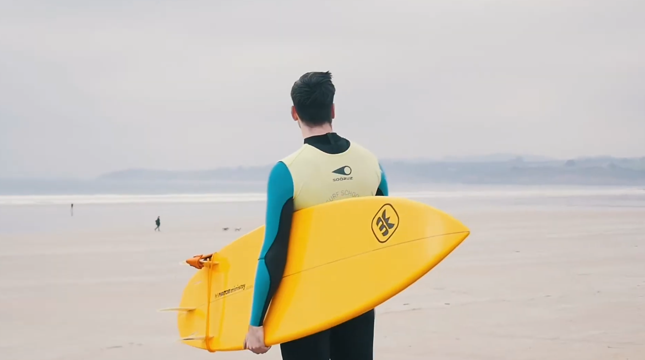Personne tenant une planche de surf jaune sur une plage, face à la mer calme sous un ciel couvert