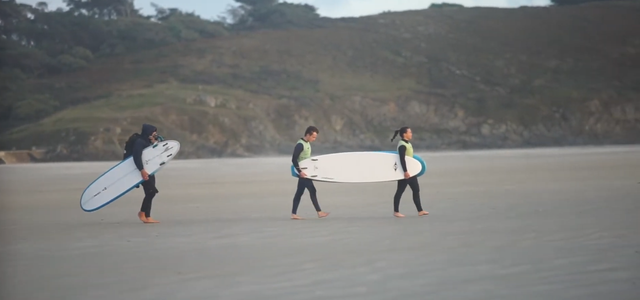 Trois surfeurs portant des planches traversent une plage, avec des collines rocheuses en arrière-plan
