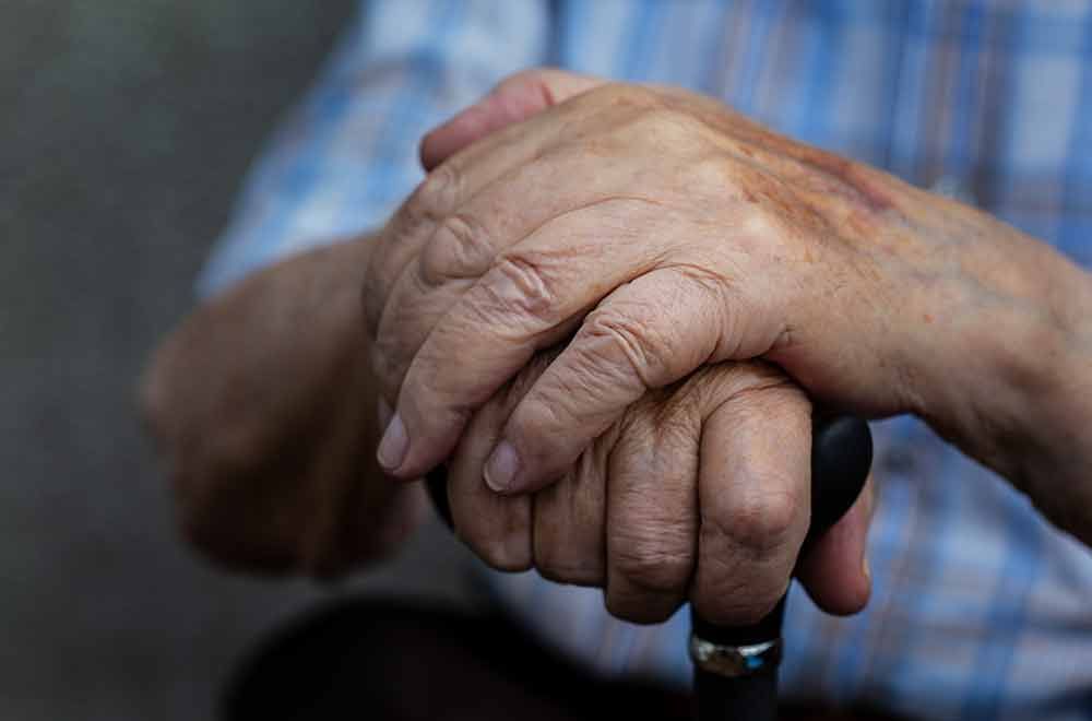 Nahaufnahme der Hände einer älteren Person, die auf einem Gehstock ruhen. Die Haut ist faltig, und der Gehstock hat einen schwarzen Griff.