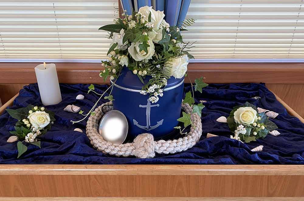 Blumenarrangement in einem blauen Eimer mit einem Seilkranz auf einem Tisch mit Blumen, einer Kerze und blauem Stoff.