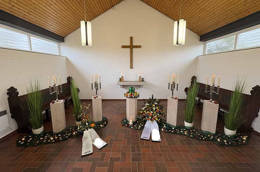 Innenansicht einer Kapelle mit Kreuz, Kerzen und Blumenarrangements.