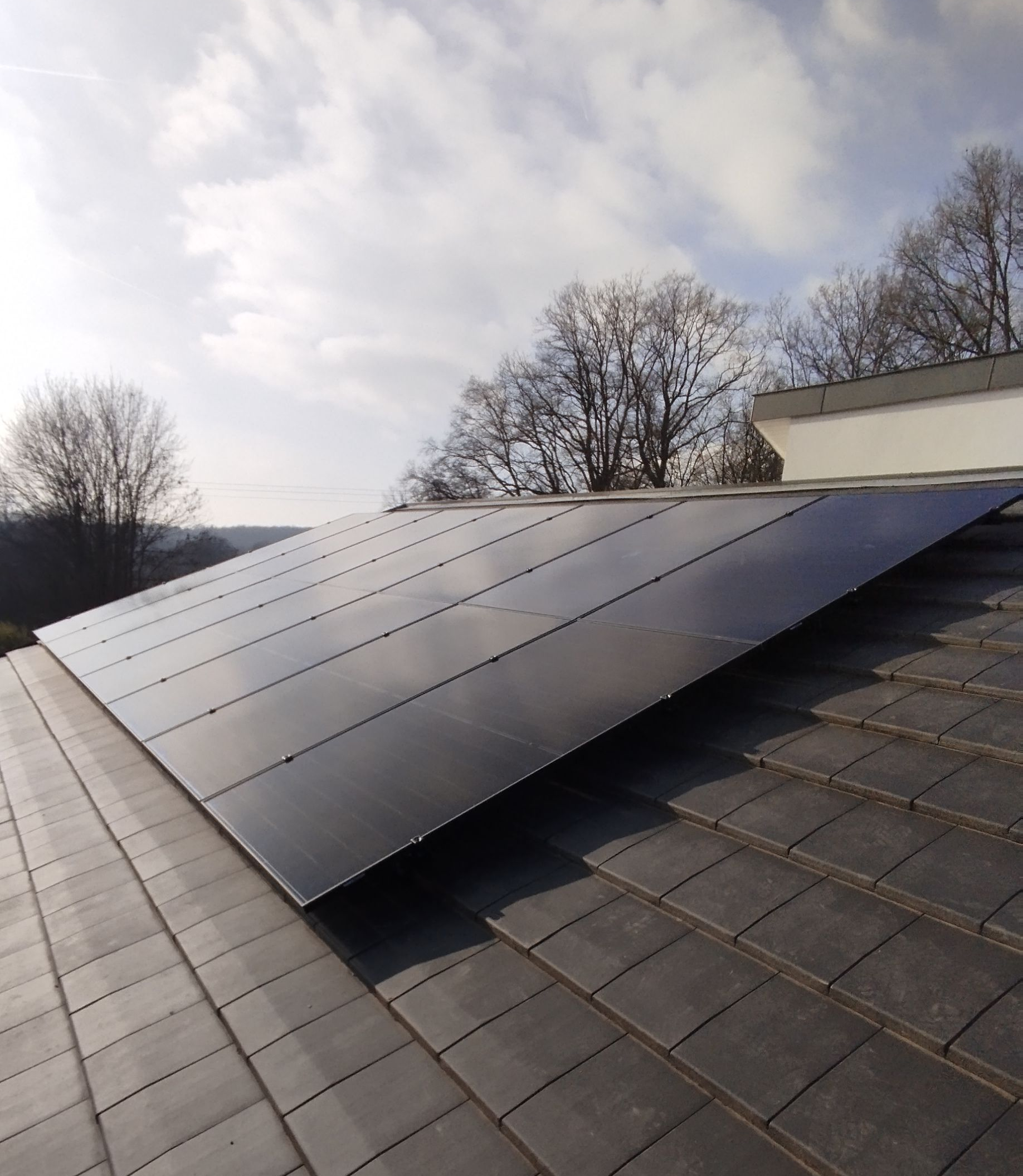 Deux personnes installent des panneaux photovoltaïques sur un toit de tuiles rouges sous un ciel nuageux.