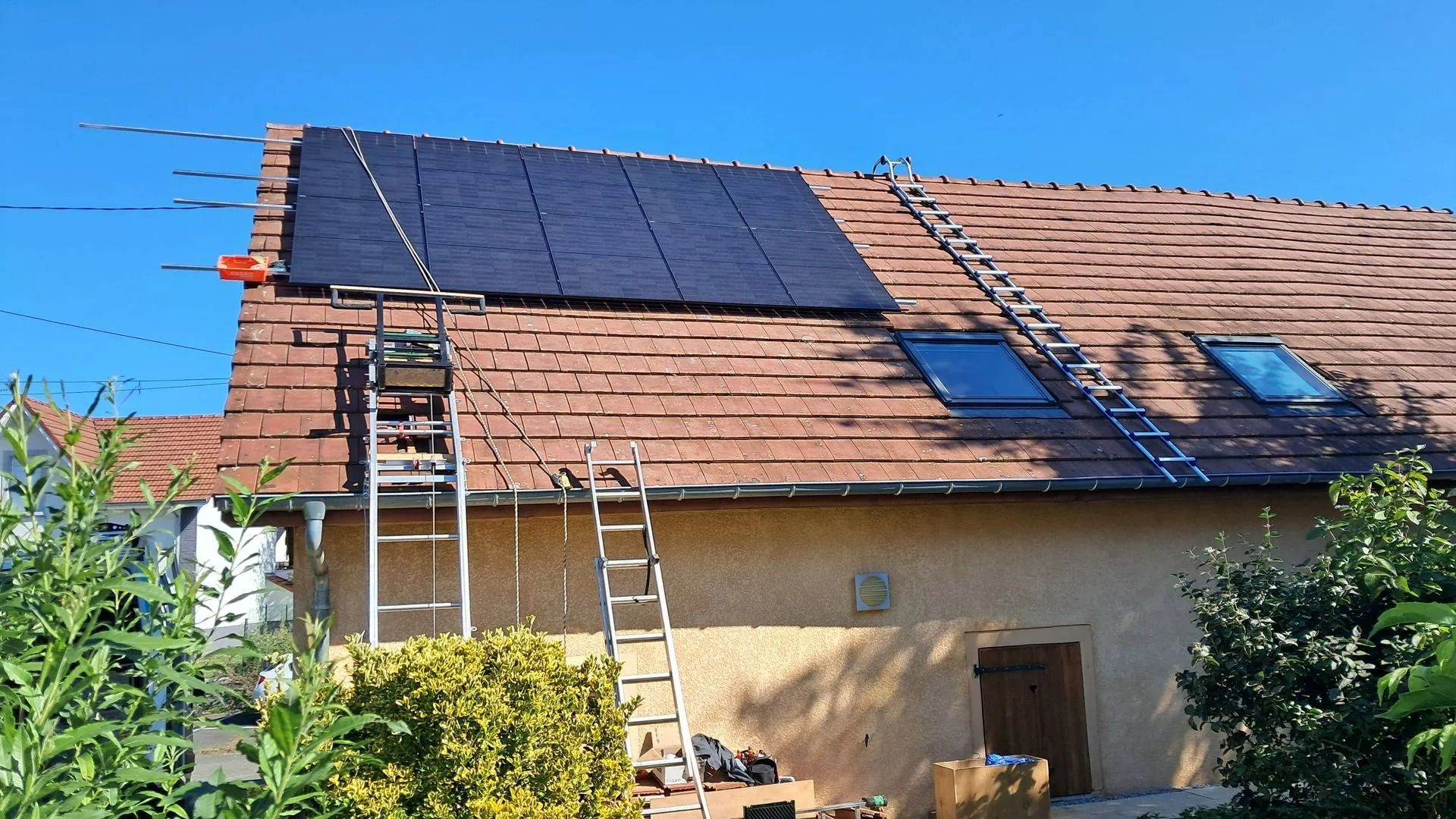 Panneaux photovoltaïques installés sur un toit de tuiles rouges, avec échelles et puits de lumière.