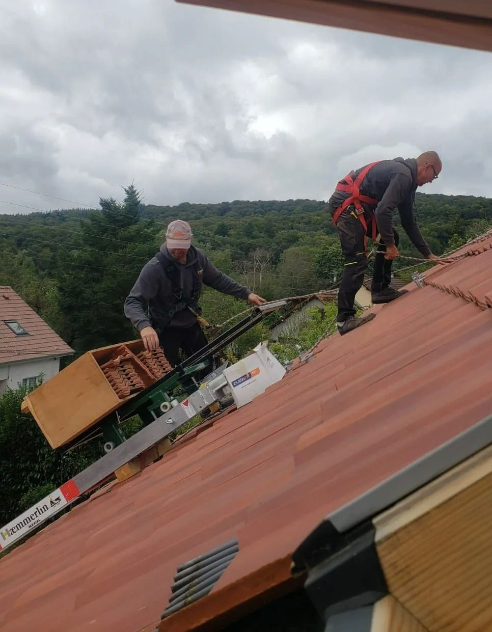 Deux couvreurs travaillent sur un toit de tuiles rouges, l'un manœuvrant une nacelle élévatrice, l'autre retenu par un harnais.