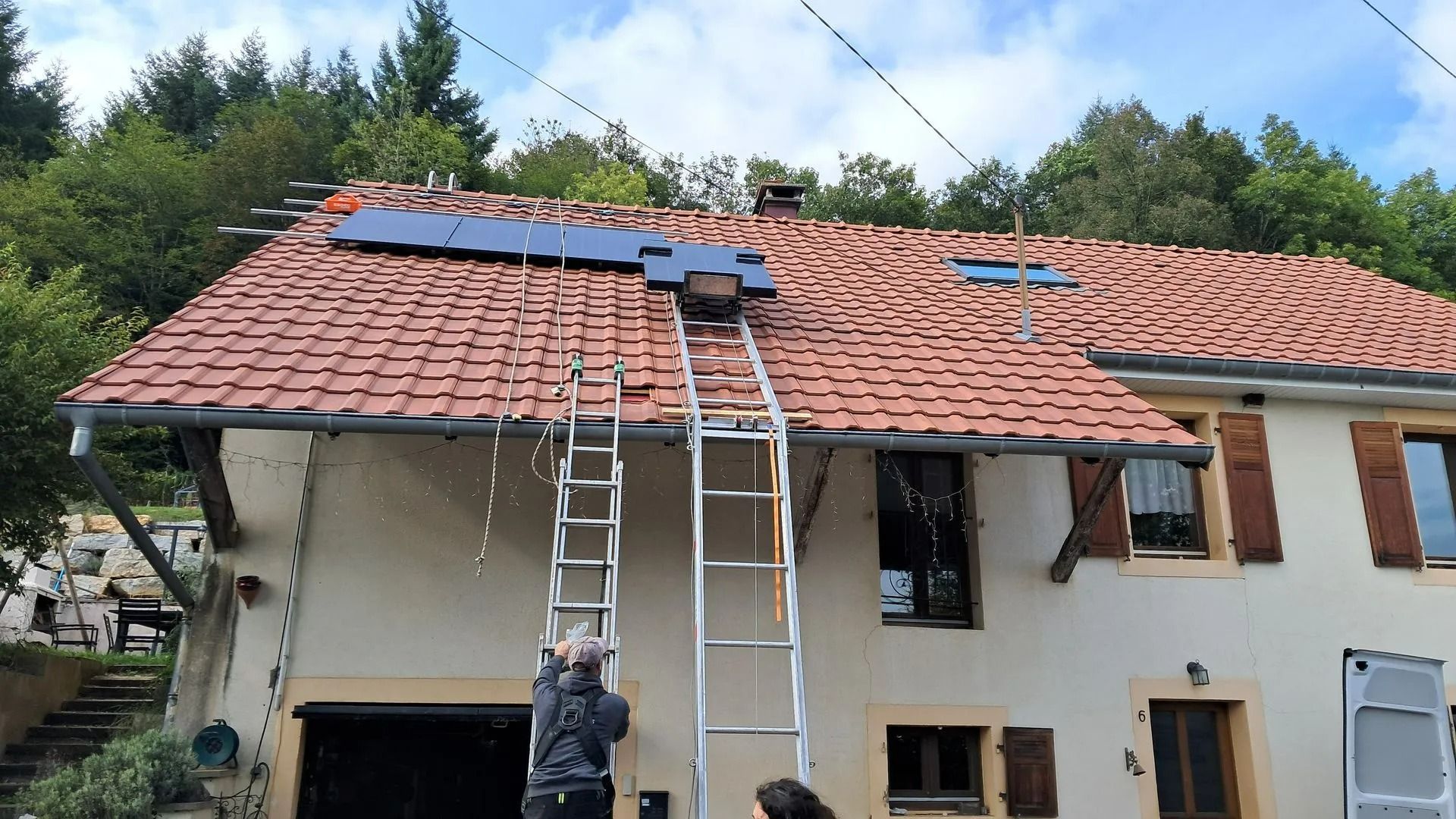 Des ouvriers installent des panneaux photovoltaïques sur le toit en tuiles rouges d'une maison claire. Ils utilisent des échelles.