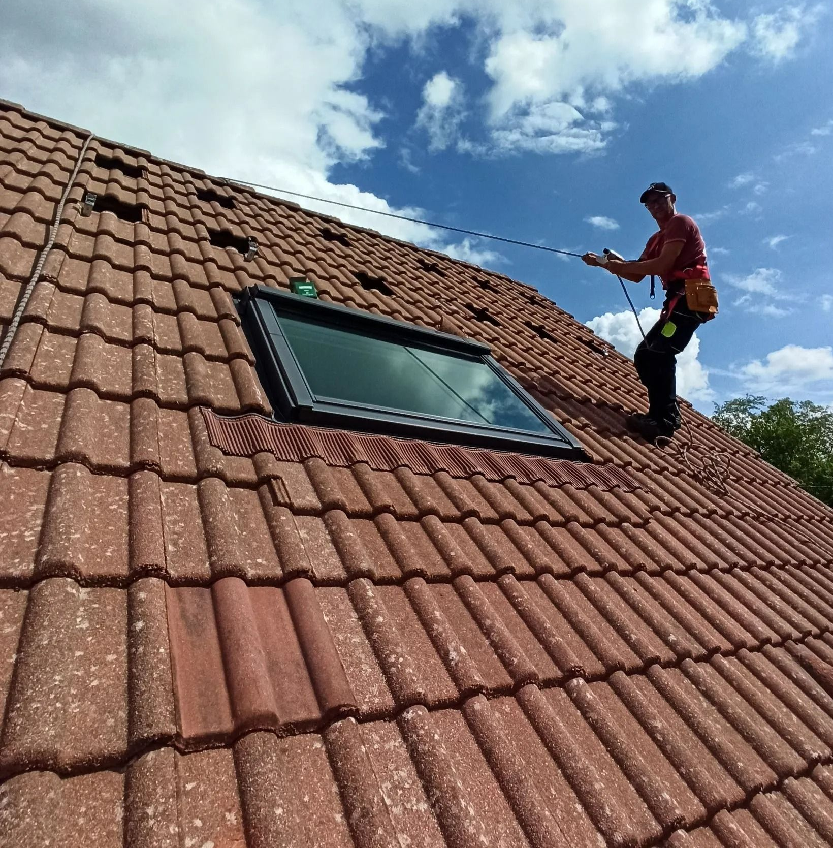 Un couvreur, harnaché et maintenu par une corde, travaille sur un toit de tuiles près d'une lucarne.