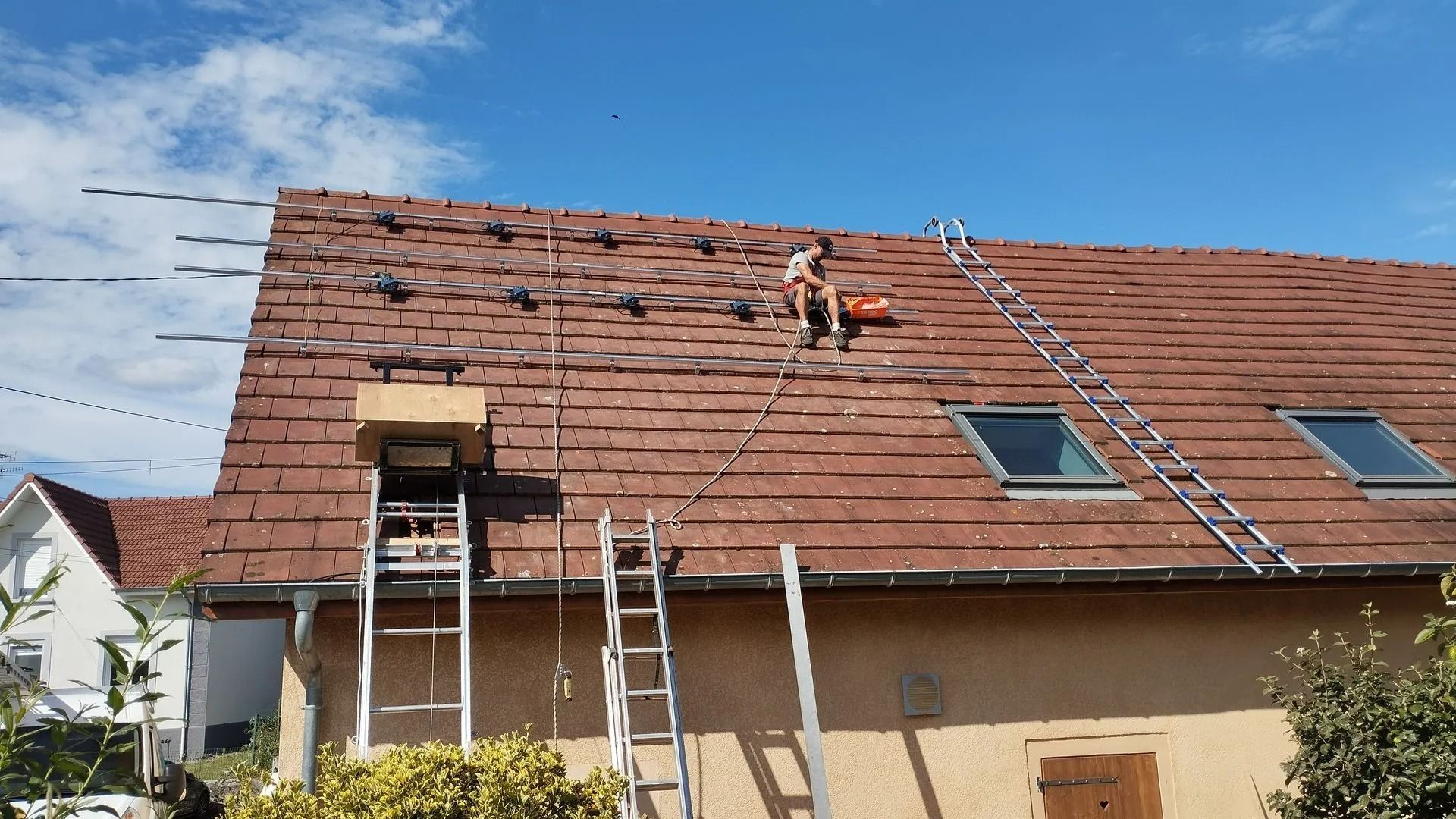 On aperçoit une personne sur un toit en train de remplacer des tuiles, deux échelles et un puits de lumière.