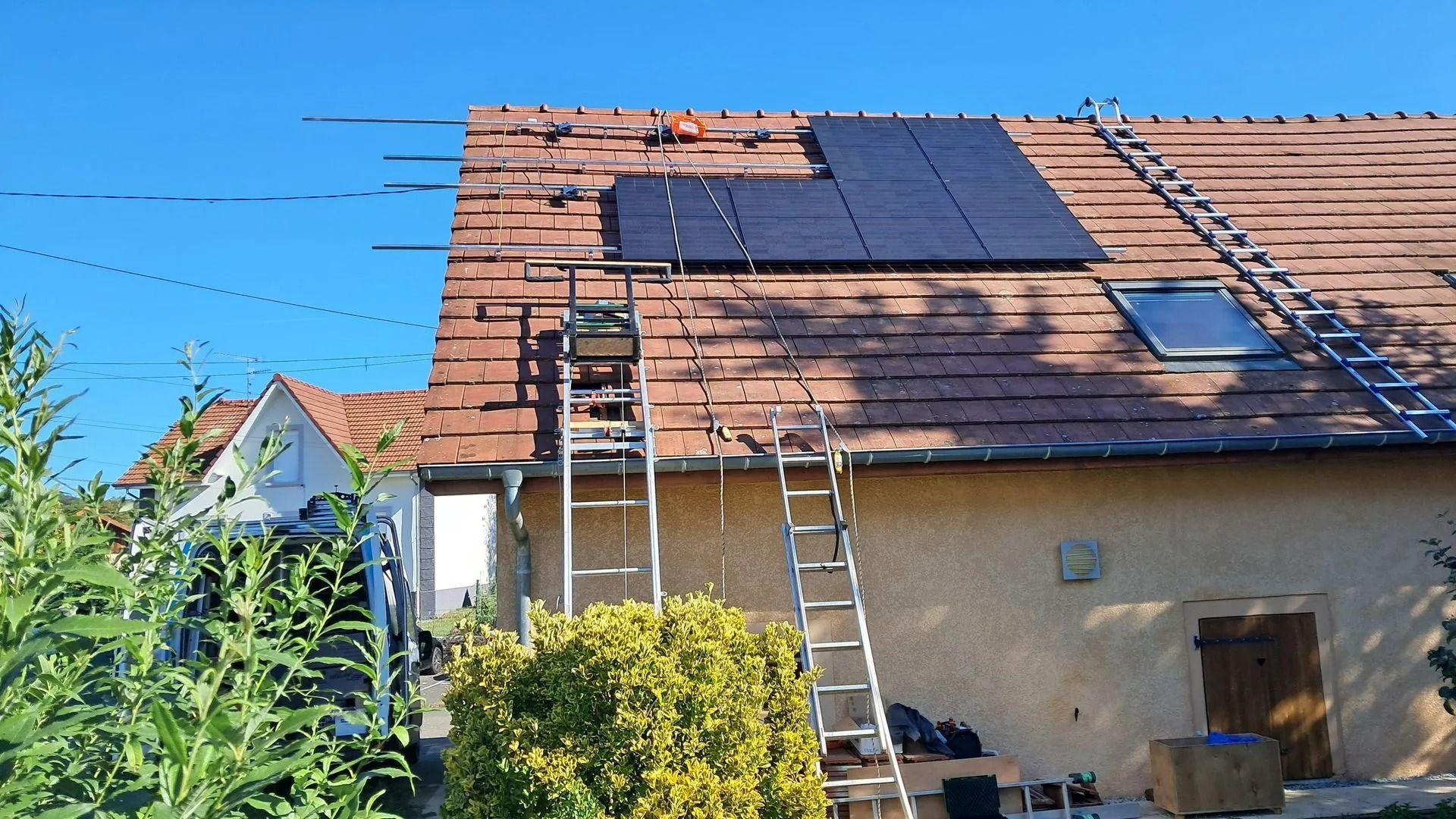 Des panneaux photovoltaïques installés sur un toit de tuiles rouges ; deux échelles appuyées contre la maison.