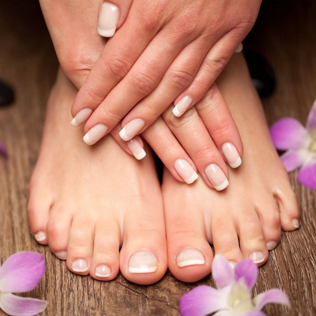 Mani con unghie curate alla francese appoggiate su piedi decorati con petali di orchidea viola su una superficie di legno.