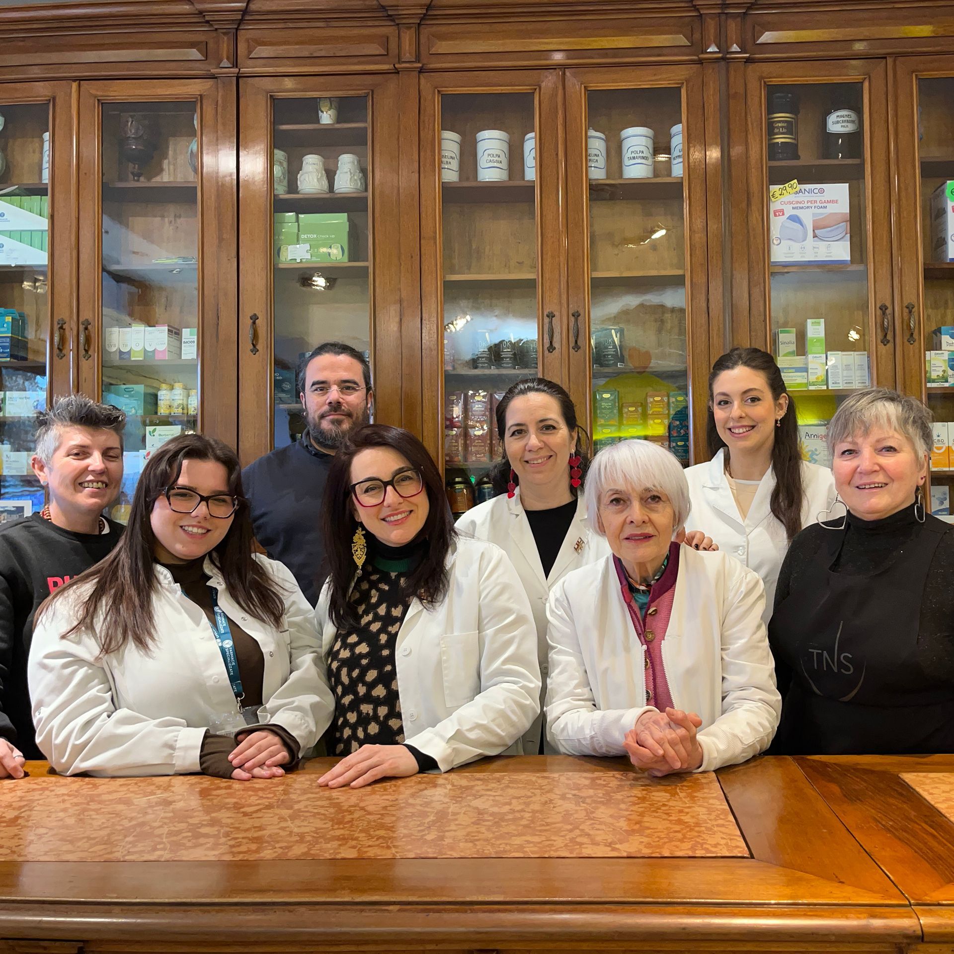Otto persone in piedi dietro un bancone di legno in una farmacia, con scaffali pieni di prodotti sullo sfondo.