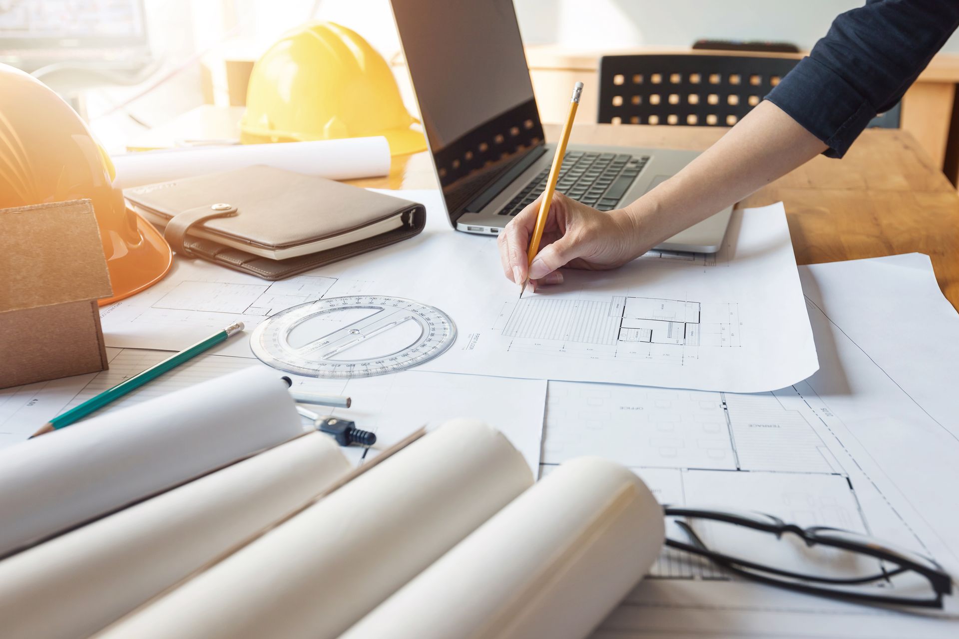 Une personne dessine des plans architecturaux à un bureau équipé d'un ordinateur portable, de plans, d'un casque de chantier et d'outils de dessin.