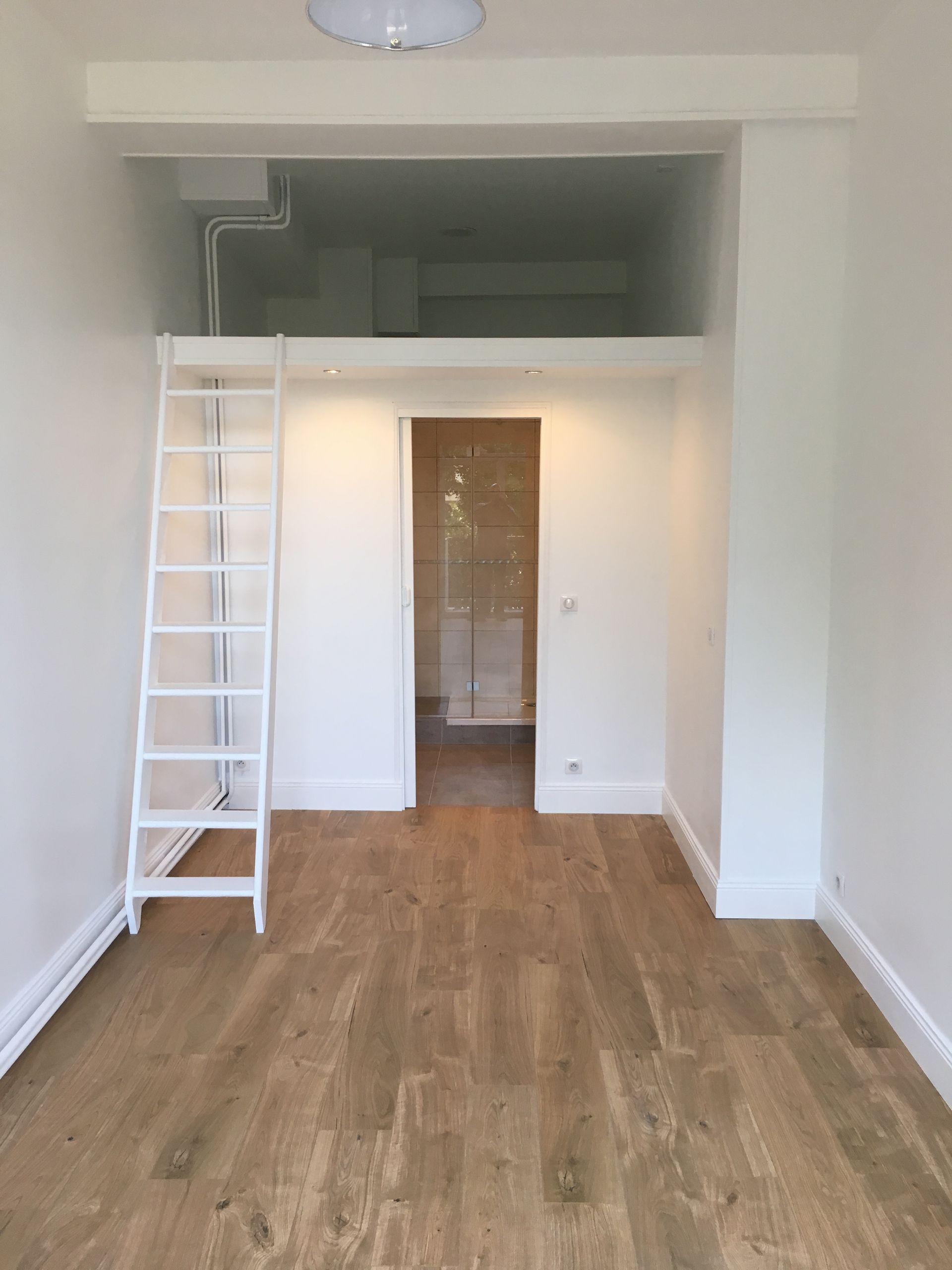 Petite chambre blanche avec un lit mezzanine et une échelle. Sol en bois clair, une porte donne sur une pièce au fond.