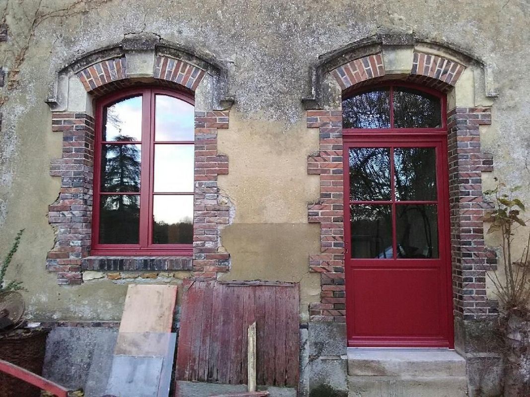 Façade d'un vieux bâtiment, avec deux fenêtres cintrées à cadre rouge et une porte rouge.