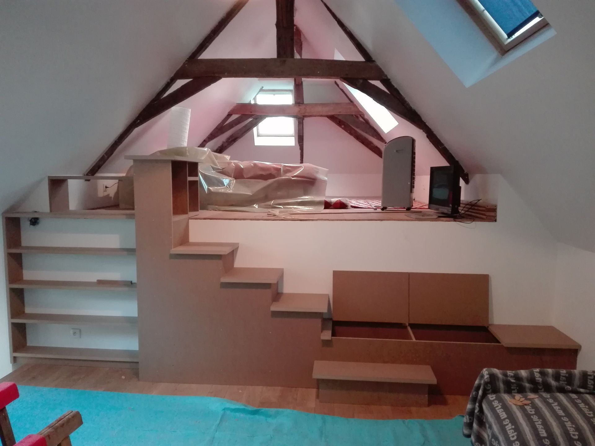 Chambre mansardée avec un lit mezzanine, des étagères blanches, un escalier en bois et une fenêtre de toit.