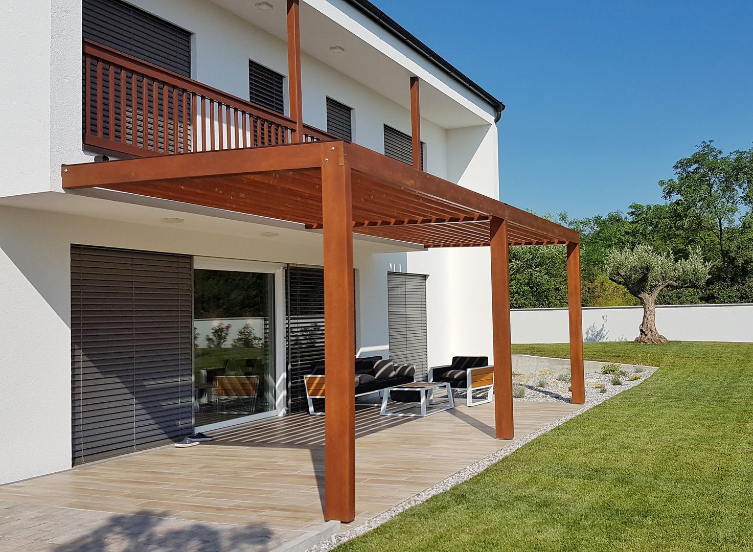 Maison blanche moderne avec une terrasse abritée par une pergola en bois, du mobilier de jardin et une pelouse sous un ciel bleu azur.