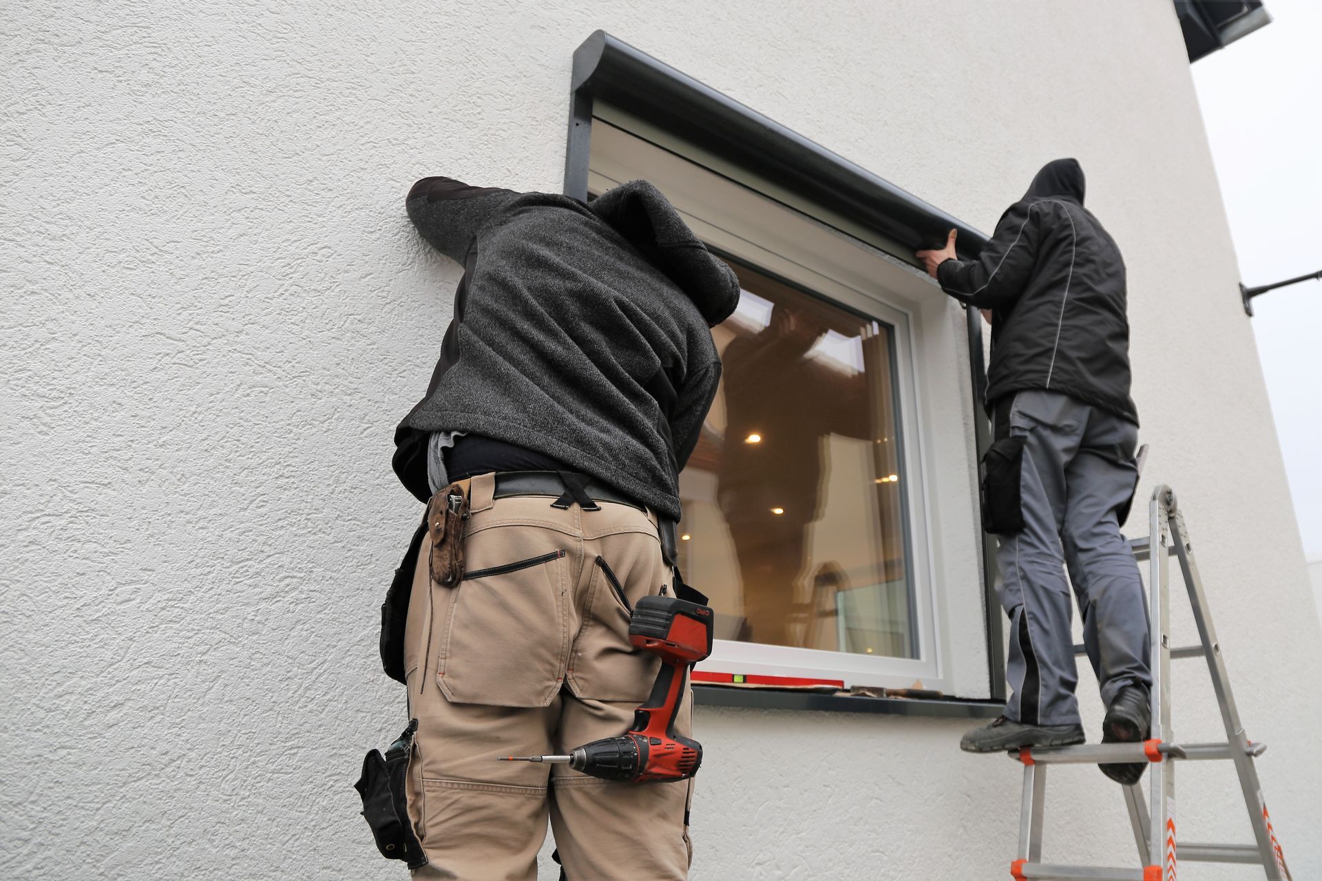 Deux ouvriers installent une fenêtre sur la façade d'une maison blanche, l'un d'eux se tenant sur une échelle.