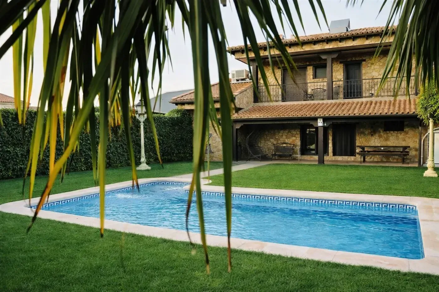 Piscina frente a casa de piedra de dos plantas con balcón y césped.