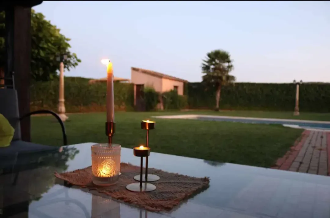 Velas sobre una mesa de cristal en un patio trasero con piscina y árboles al fondo.