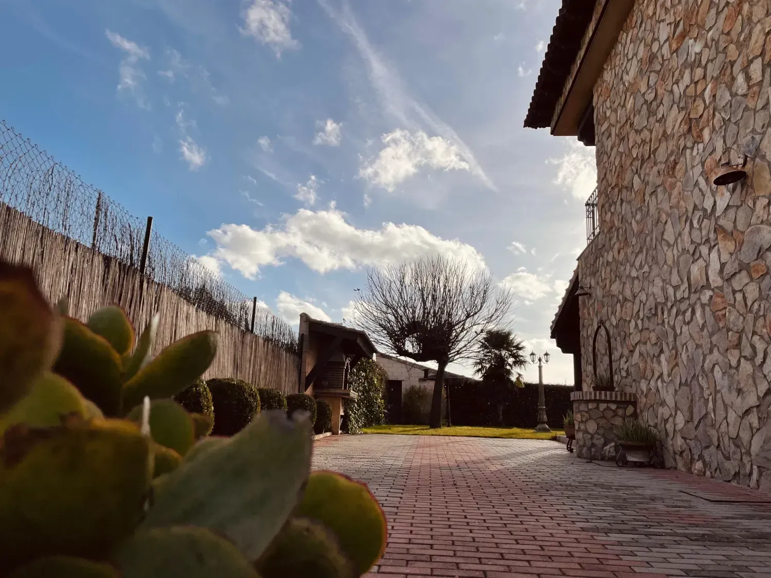 Un edificio de piedra y un patio trasero con un cielo nublado, un camino de ladrillos y cactus en primer plano.