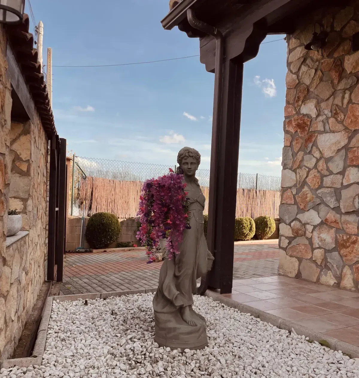 La estatua de una mujer cubierta de flores violetas se encuentra en un patio con paredes de piedra, grava y un cielo azul.