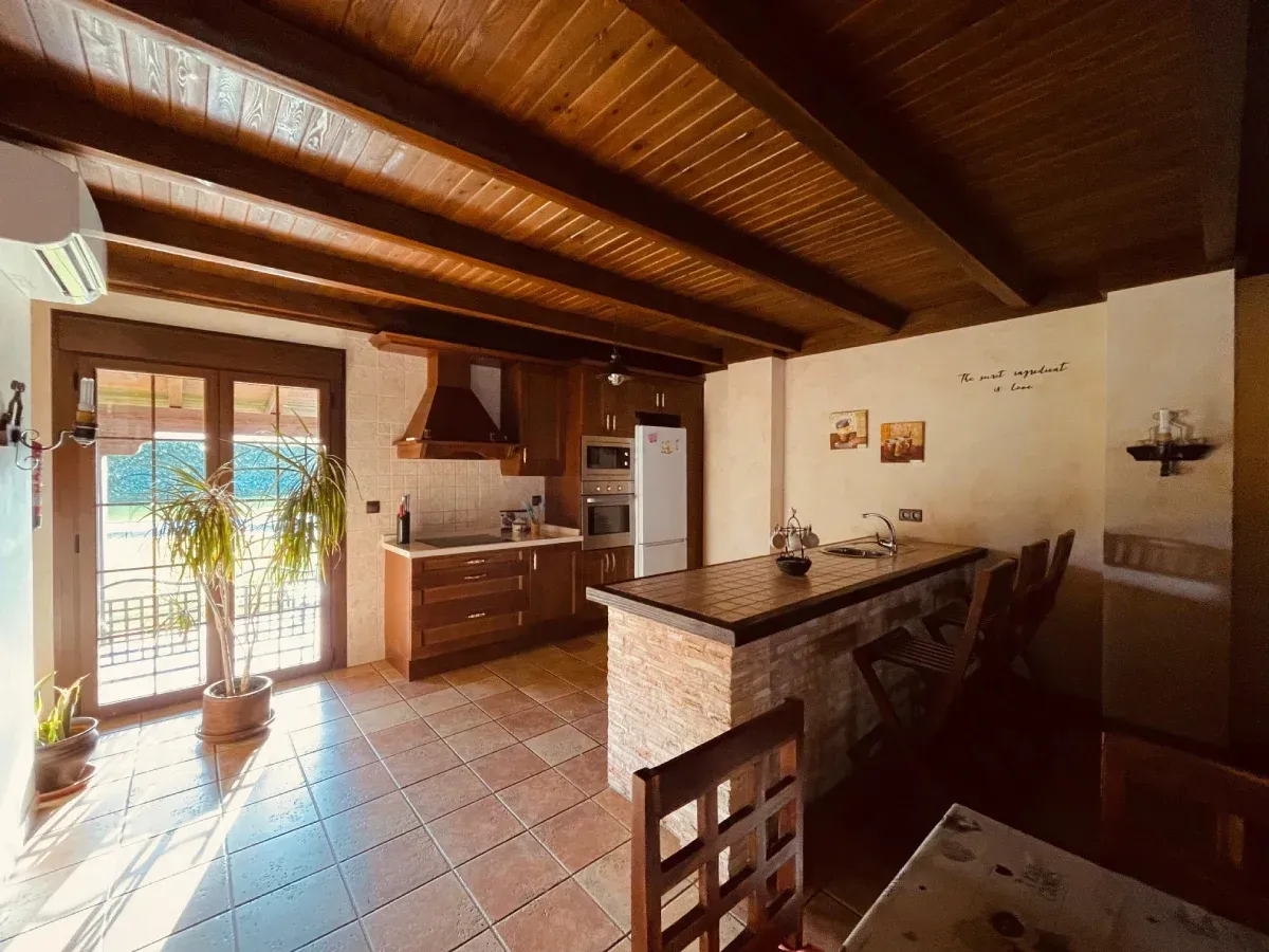Cocina con techo de madera, gabinetes e isla; una puerta de vidrio se abre a la vegetación exterior.