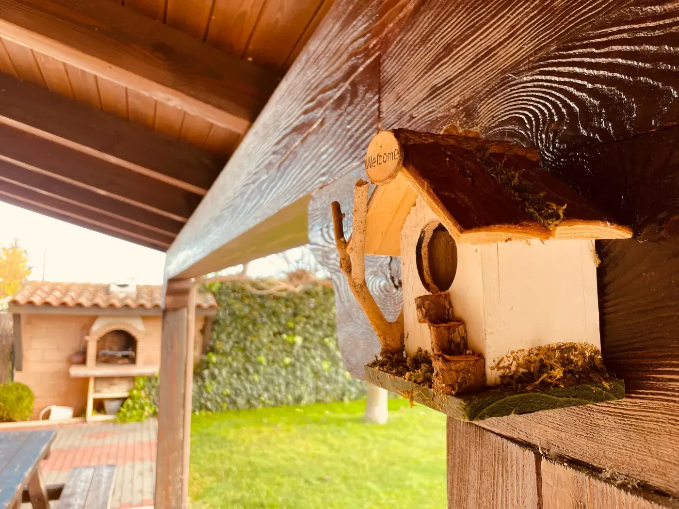 Casa de pájaros colgada de una viga de madera, patio trasero con césped verde y un pequeño horno al fondo.