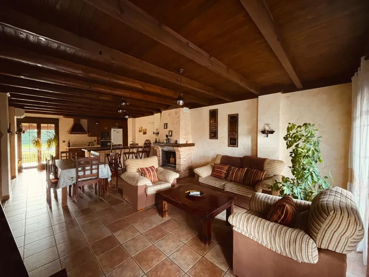 Acogedora sala de estar con chimenea, sofás y comedor. Vigas de madera, iluminación cálida y suelo de baldosas.