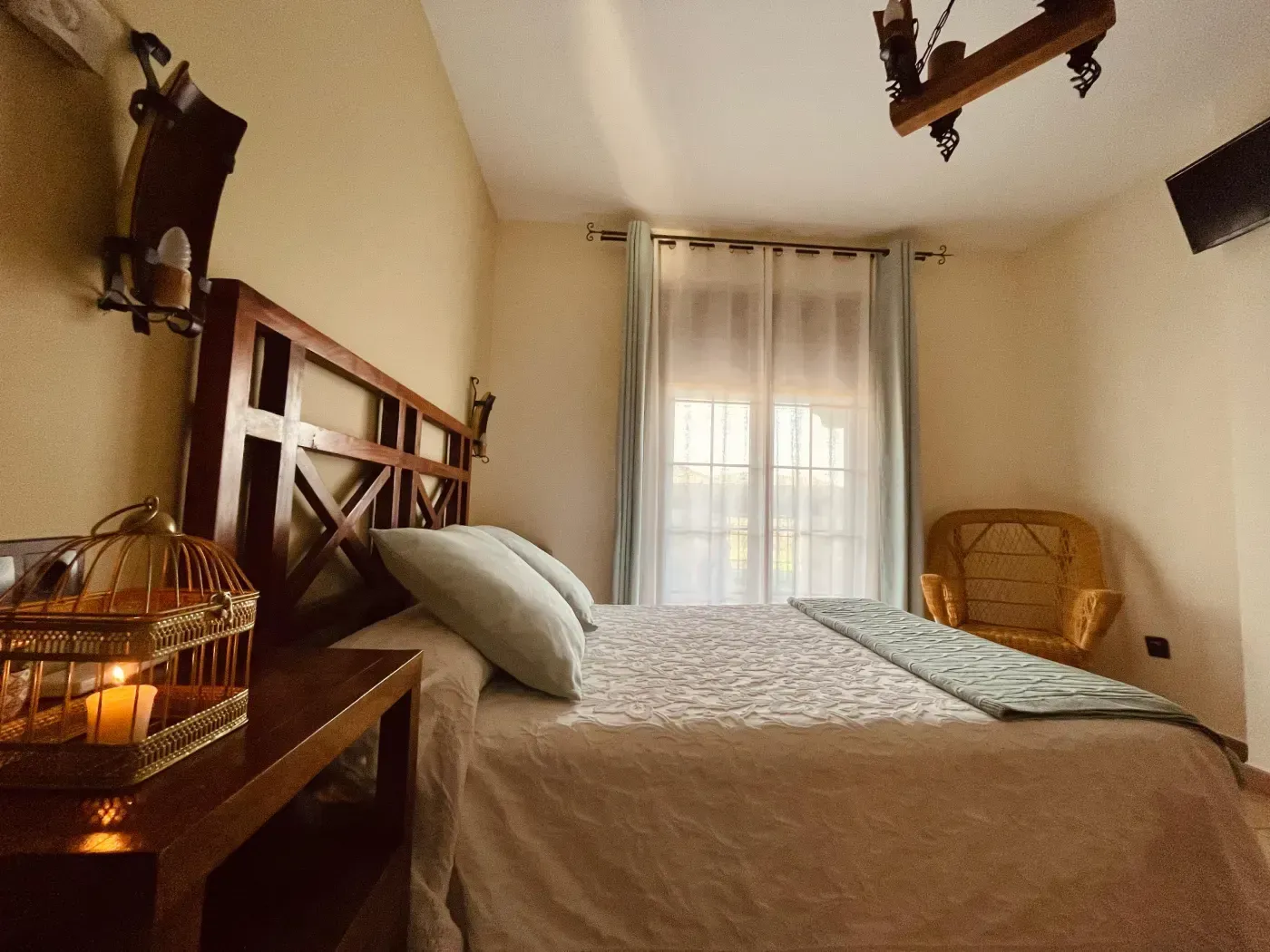 Dormitorio con cabecero de madera, mesita de noche con jaula de pájaros, cama, ventana con cortinas y silla de mimbre.