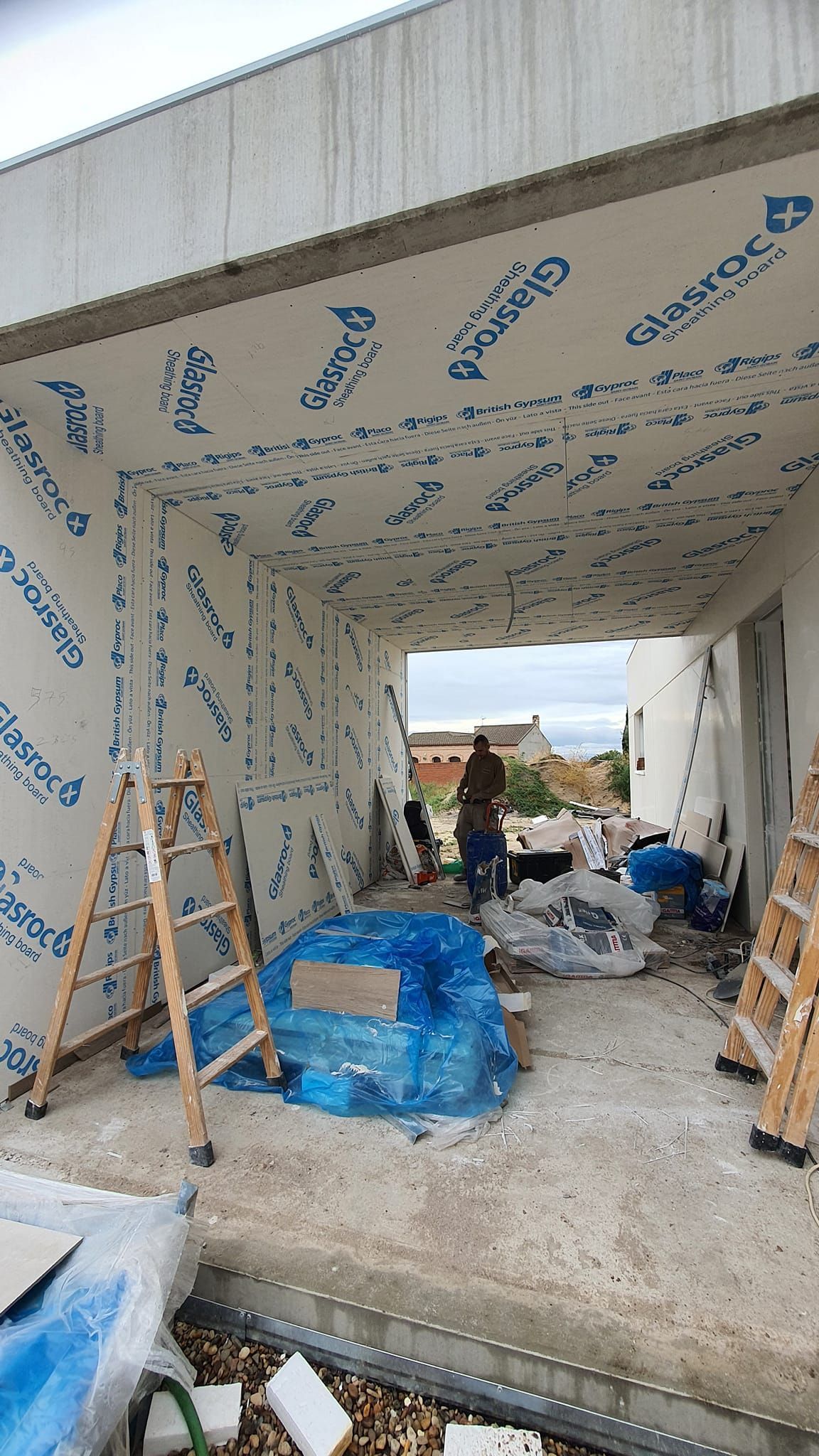 Interior de una obra en construcción: una persona entre materiales, paredes cubiertas con aislamiento plano, escaleras y una puerta abierta.