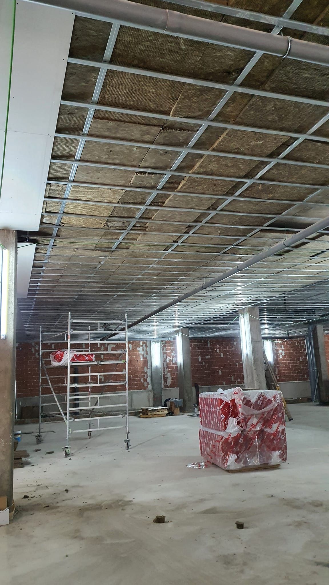 Interior de una obra con techo de rejilla parcialmente instalado sobre una pared de ladrillo. Hay andamios y materiales.