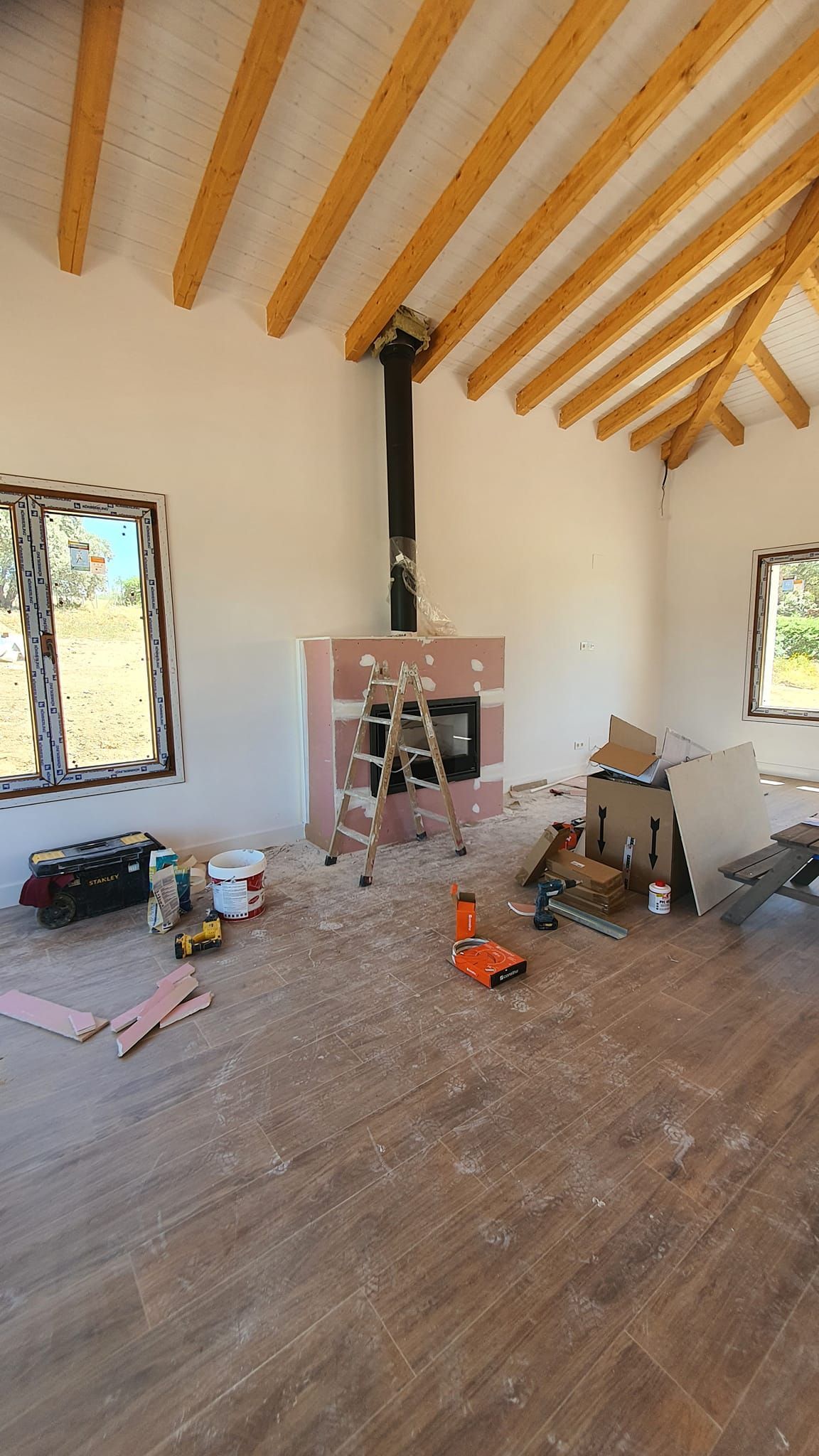 Interior de una habitación en construcción con vigas a la vista, chimenea, ventanas, herramientas y materiales de construcción.