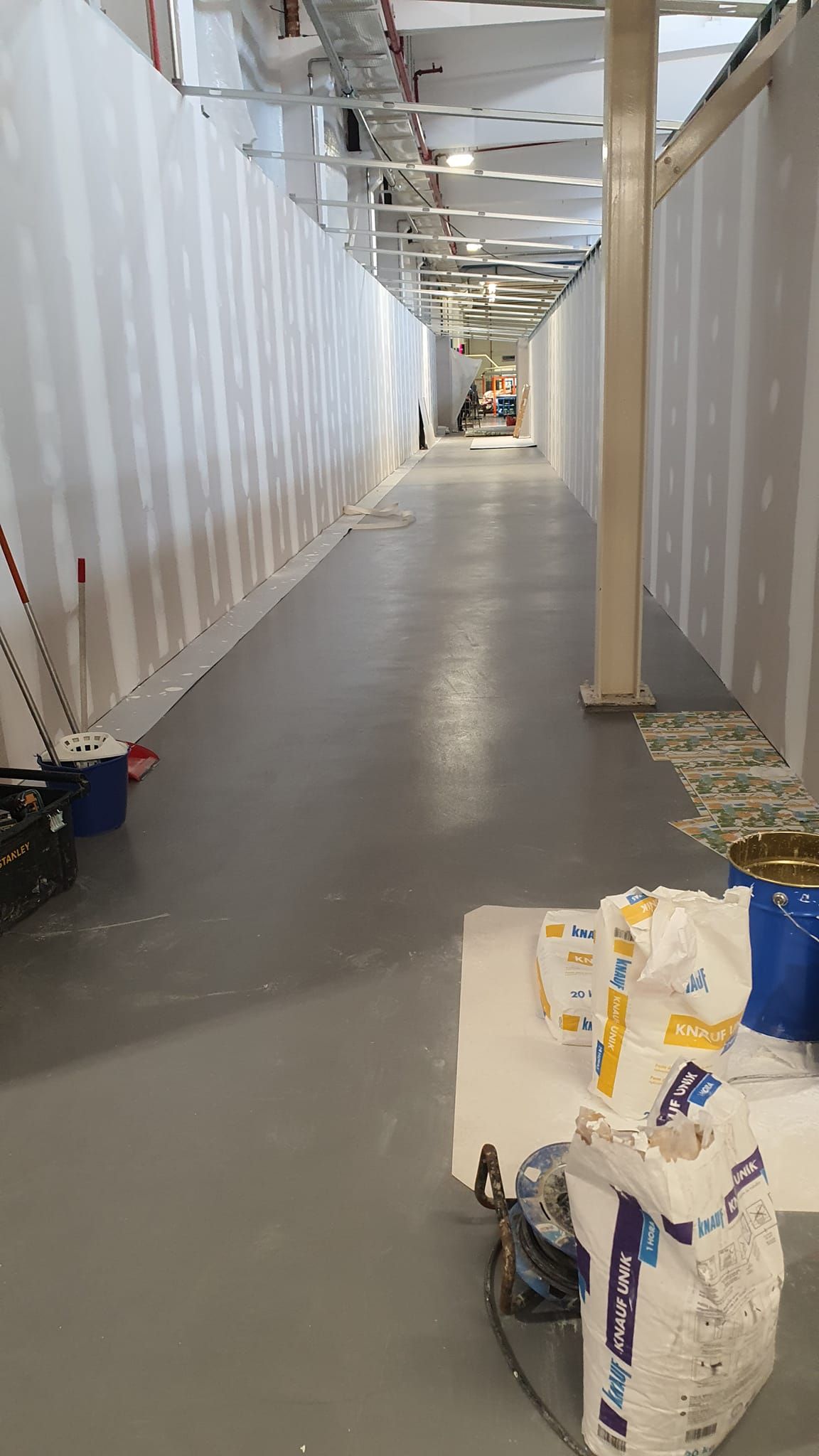 Pasillo largo y estrecho en construcción con piso gris, paredes blancas y materiales de construcción.