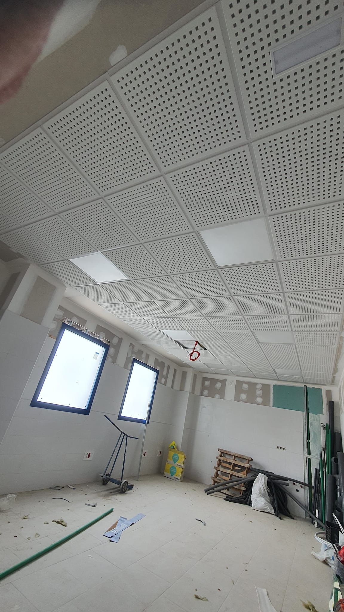 Habitación interior con techo perforado blanco, ventanas y materiales de construcción.