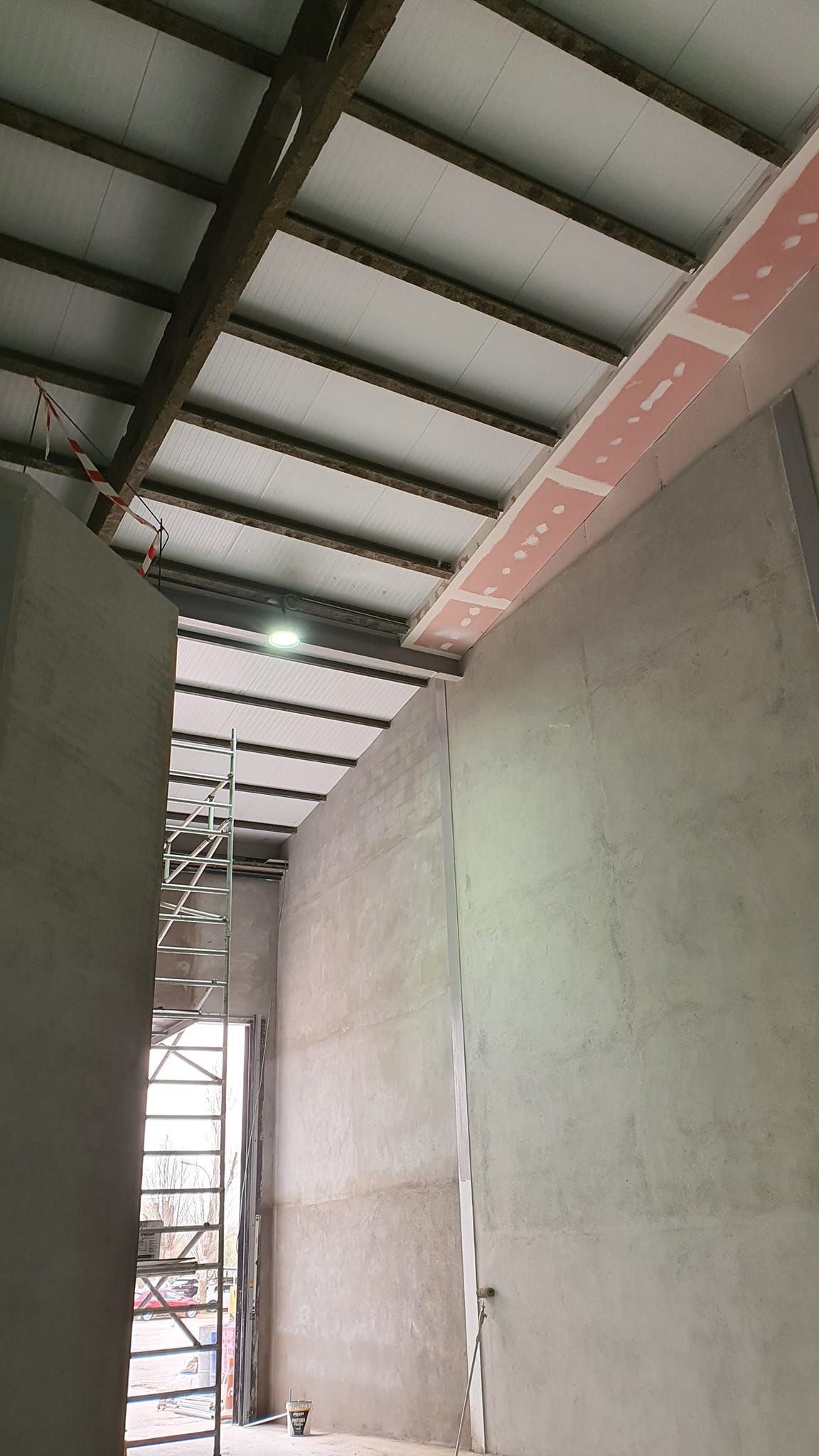 Interior de una obra en construcción: paredes grises, techo de metal, andamios, una franja rosa y una luz.
