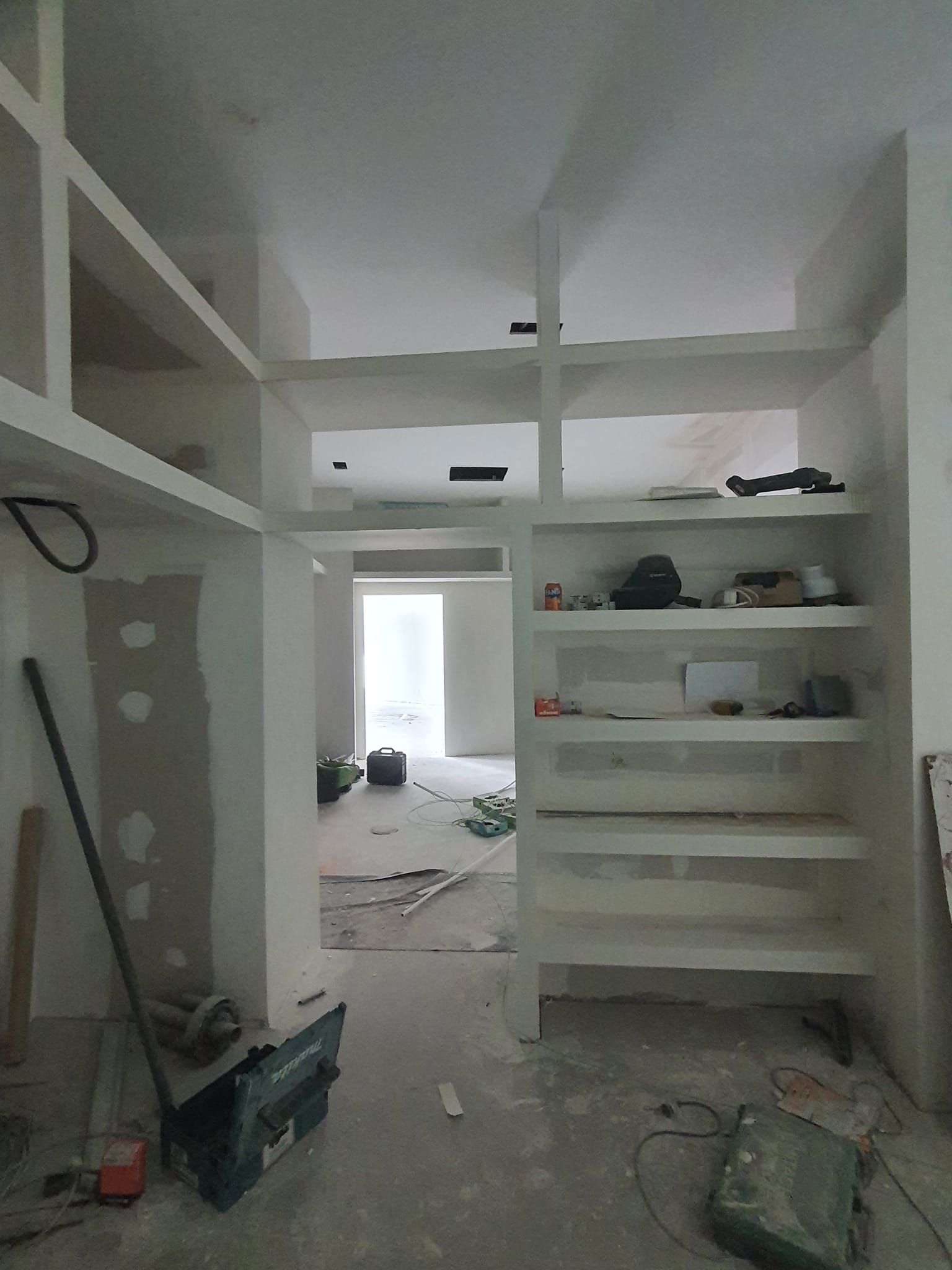 Interior en construcción: estanterías en construcción. Puerta abierta al fondo. Superficies blancas.