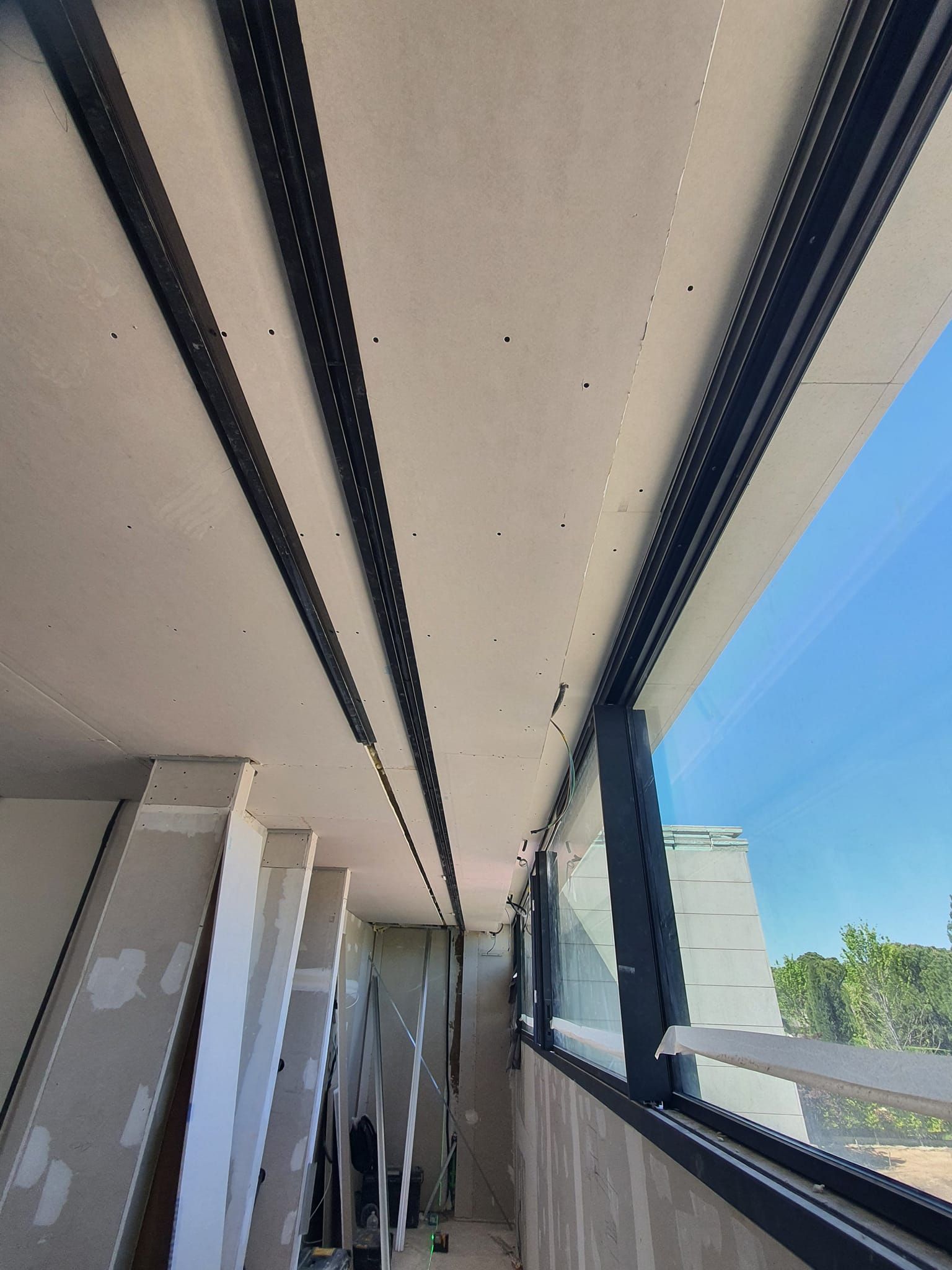 Vista interior de un sitio de construcción, con un techo con vigas oscuras, ventanas grandes y pilas de paneles de yeso.