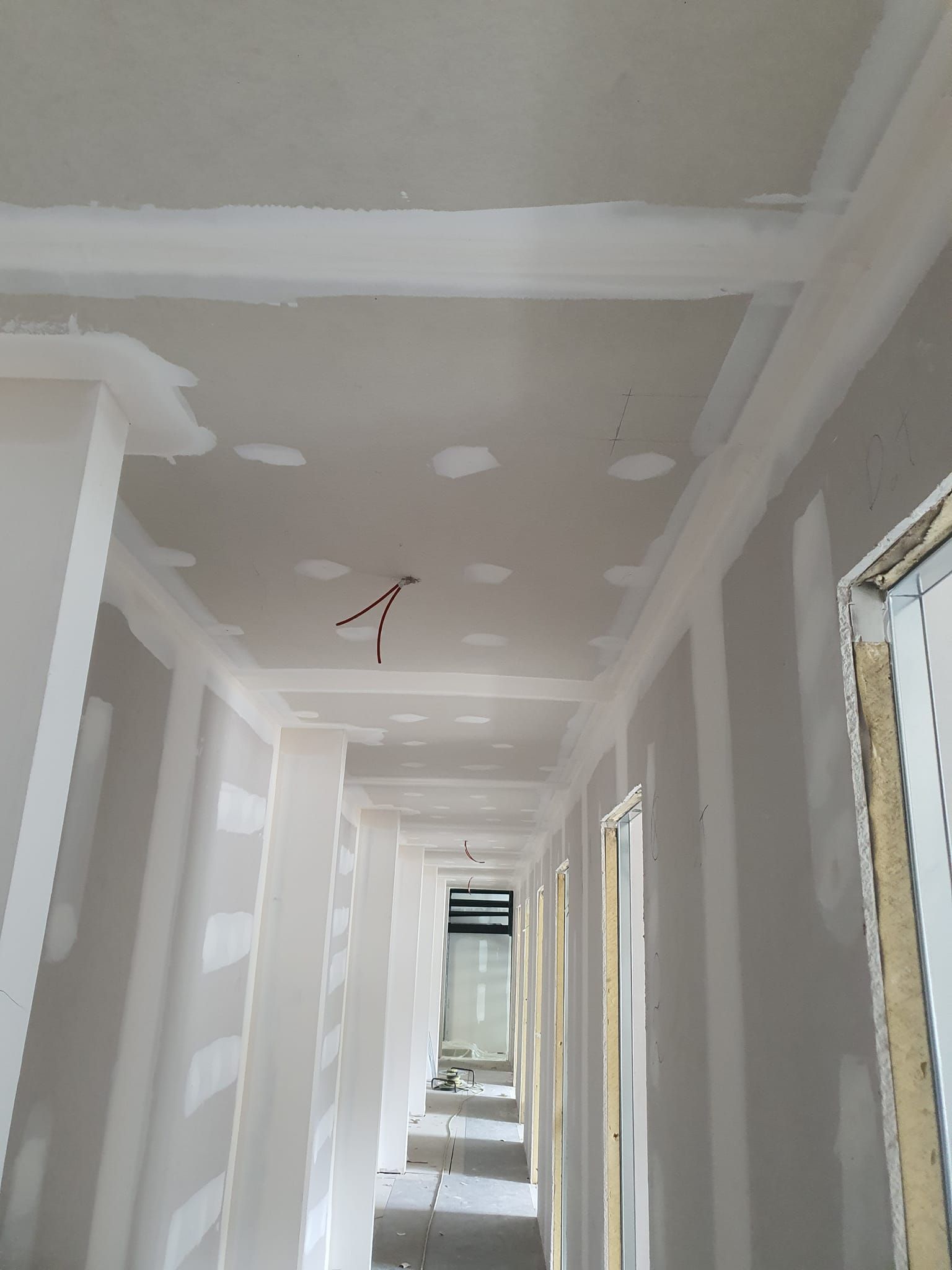 Un pasillo en construcción: paneles de yeso en paredes y techo, con cables eléctricos colgando.