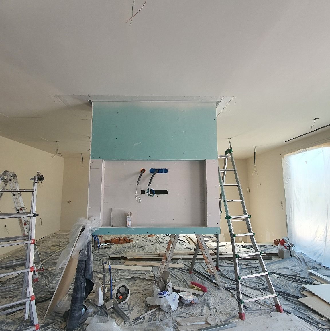 Habitación en construcción; paneles de yeso en la pared con accesorios de plomería, escaleras, herramientas y escombros en el piso.