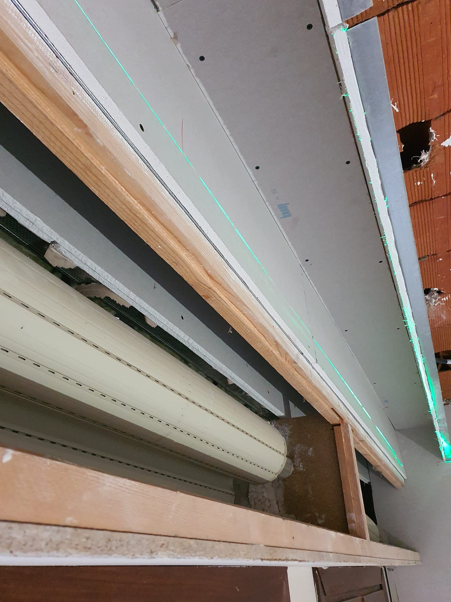 Obra con nivel láser verde en paneles de yeso y tuberías expuestas. Estructura de madera y ladrillo visibles.