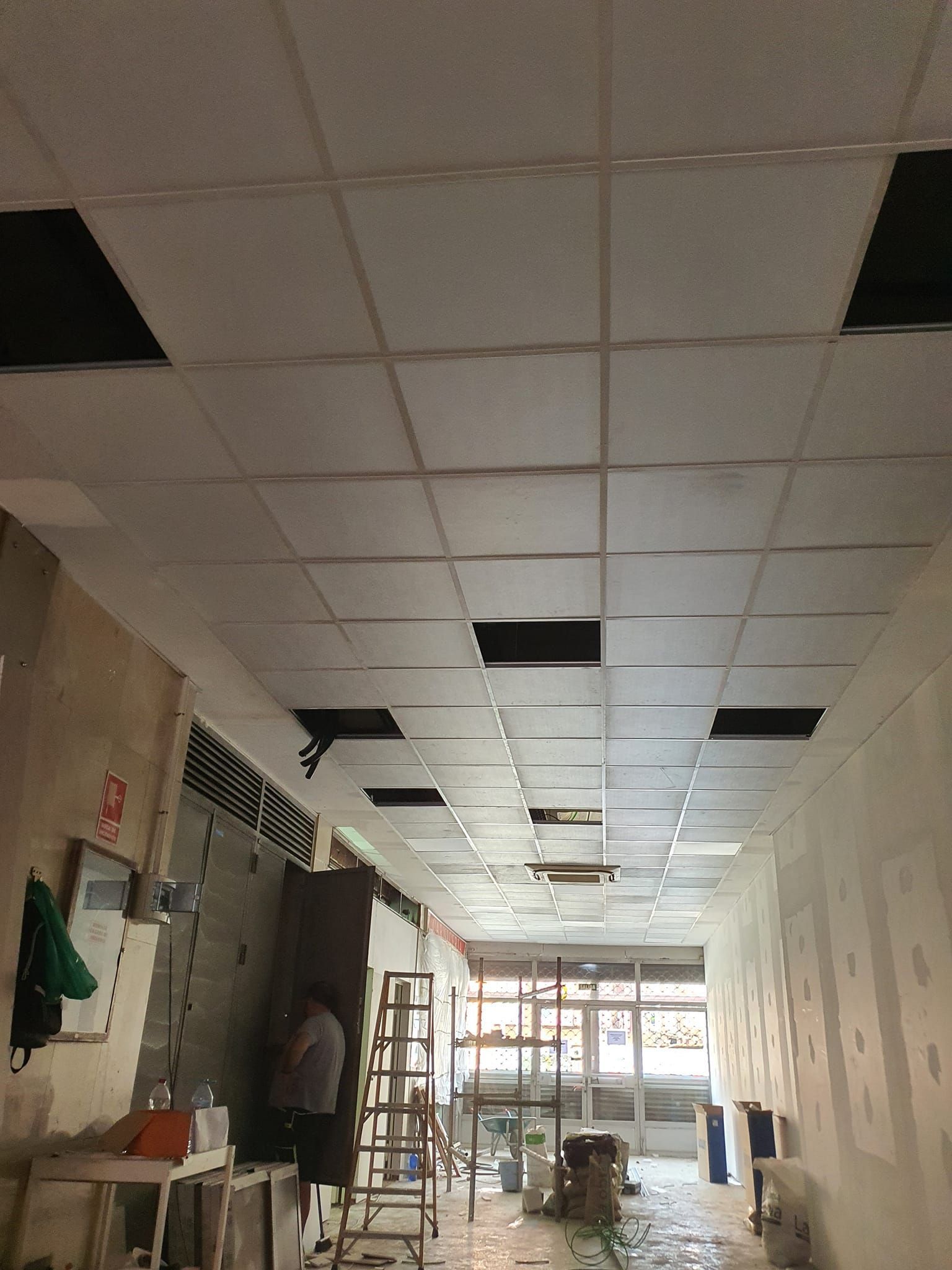 Un techo al que le faltan tejas, revelando un espacio negro. Una persona trabaja en una escalera en un pasillo blanco.