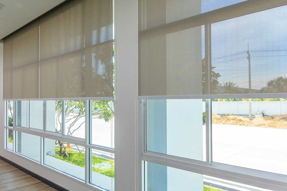 Des fenêtres de bureau baignées de soleil, avec des stores beiges, donnent sur une allée extérieure lumineuse.