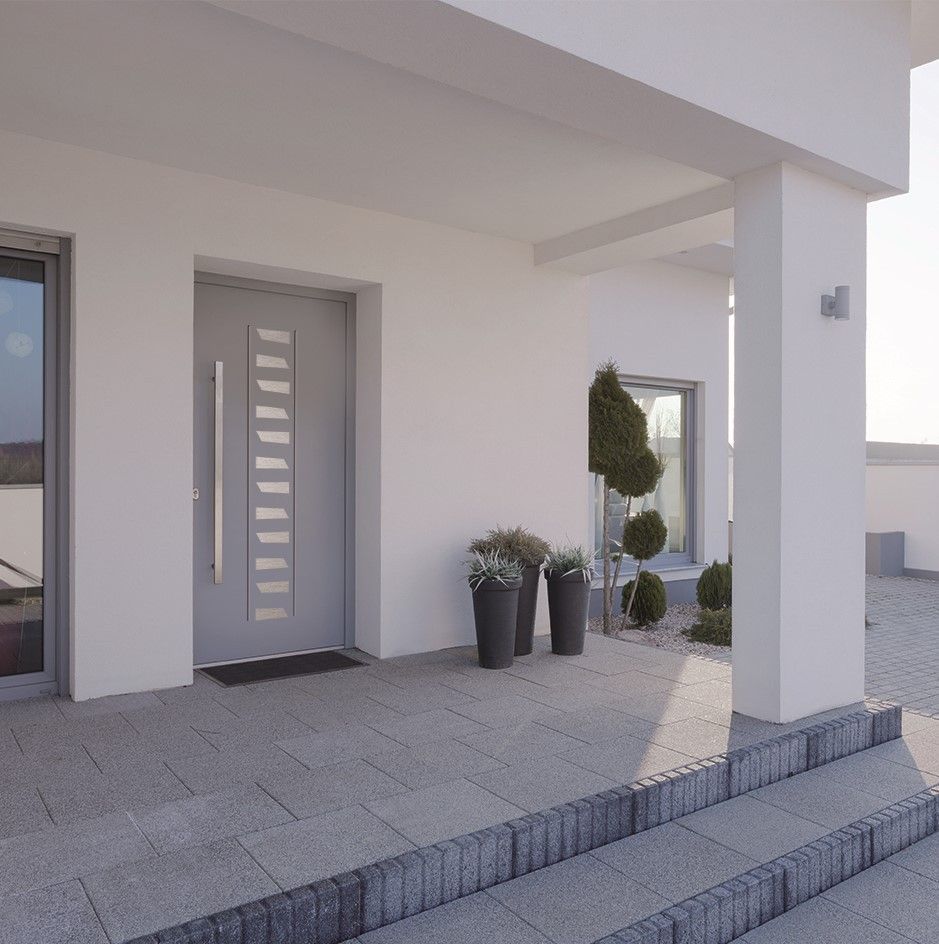 Entrée d'une maison moderne blanche avec une porte d'entrée grise, des plantes en pot et des marches carrelées sous un porche couvert.