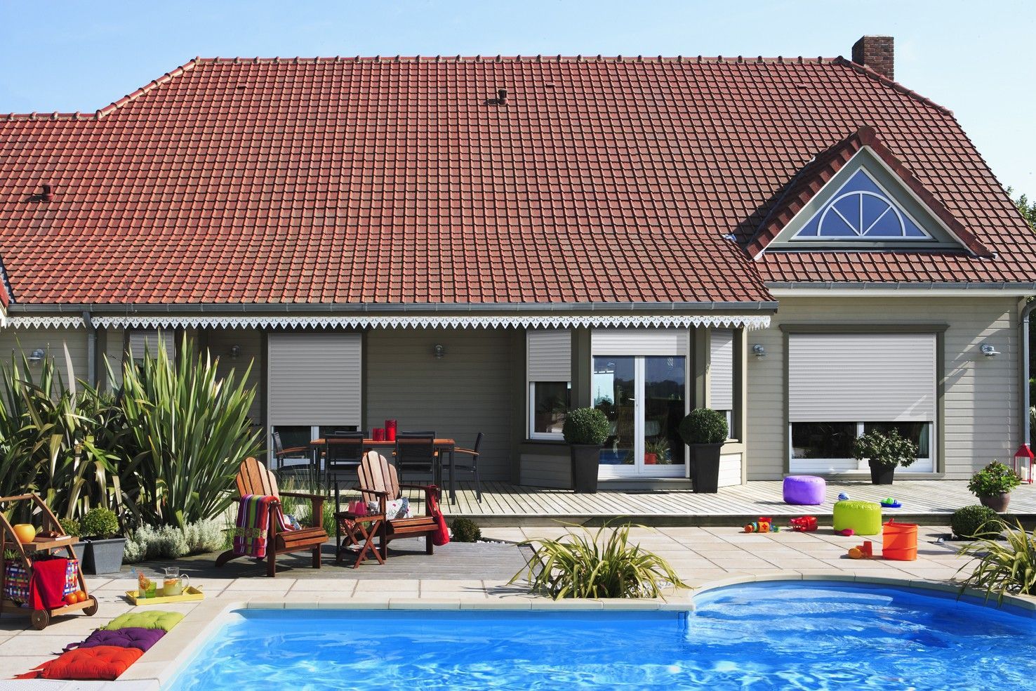 Jardin avec une maison, des meubles de terrasse et une piscine bleue sous un toit de tuiles rouges.