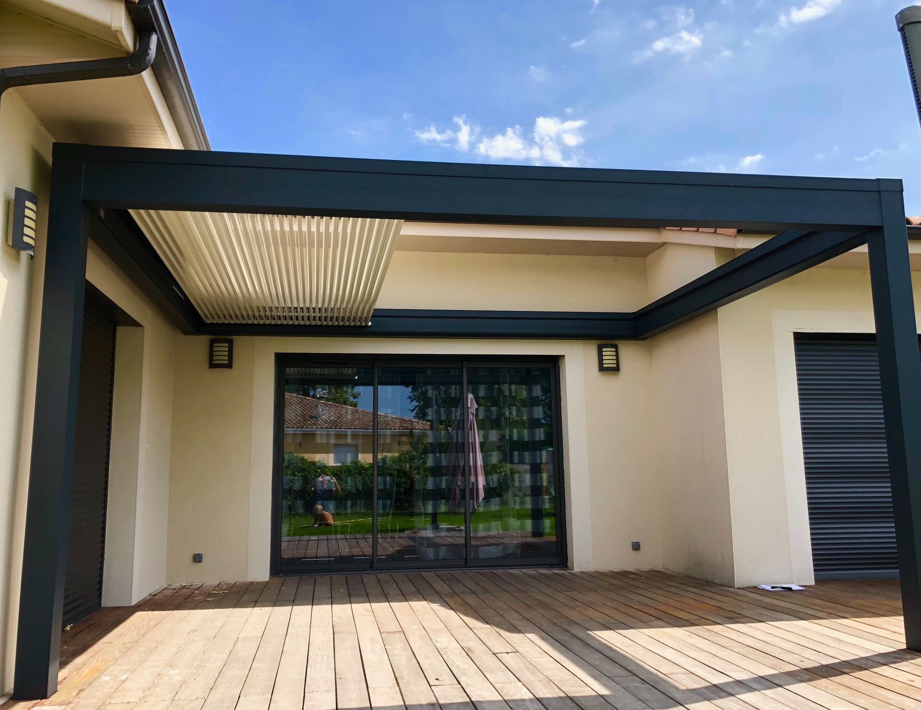 Patio moderne avec pergola en métal foncé, portes coulissantes en verre et terrasse en bois devant une maison beige