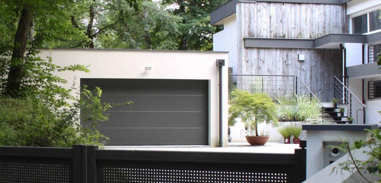 Maison blanche moderne avec garage gris, balcon et plantes en pot derrière une clôture noire