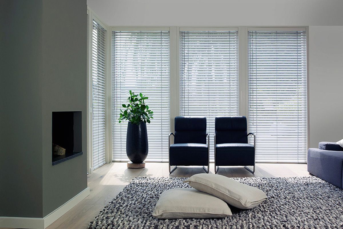 Salon moderne avec stores blancs, chaises bleues, plante en pot, cheminée et tapis gris avec coussins.