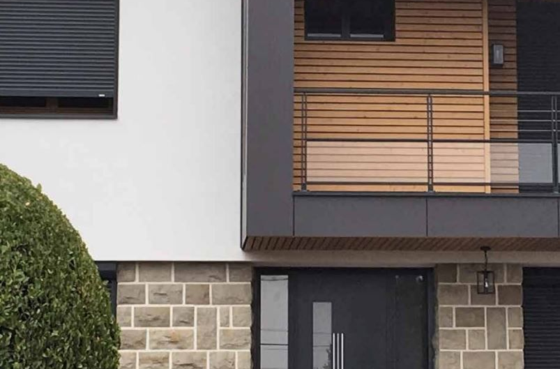 Façade de maison moderne avec soubassement en pierre, murs blancs, boiseries foncées et volets de balcon en bois.